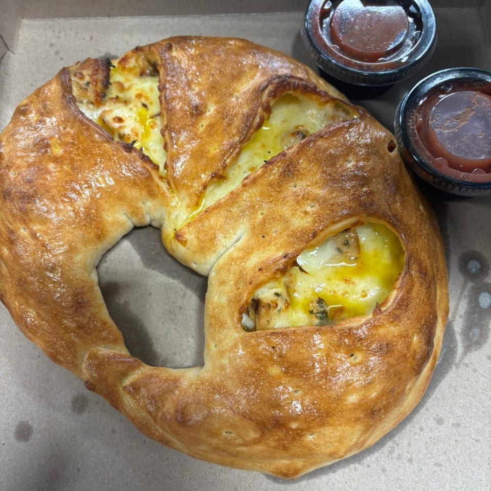 Golden-brown pizza pretzel with melted cheese and two dipping sauces in a takeout box.