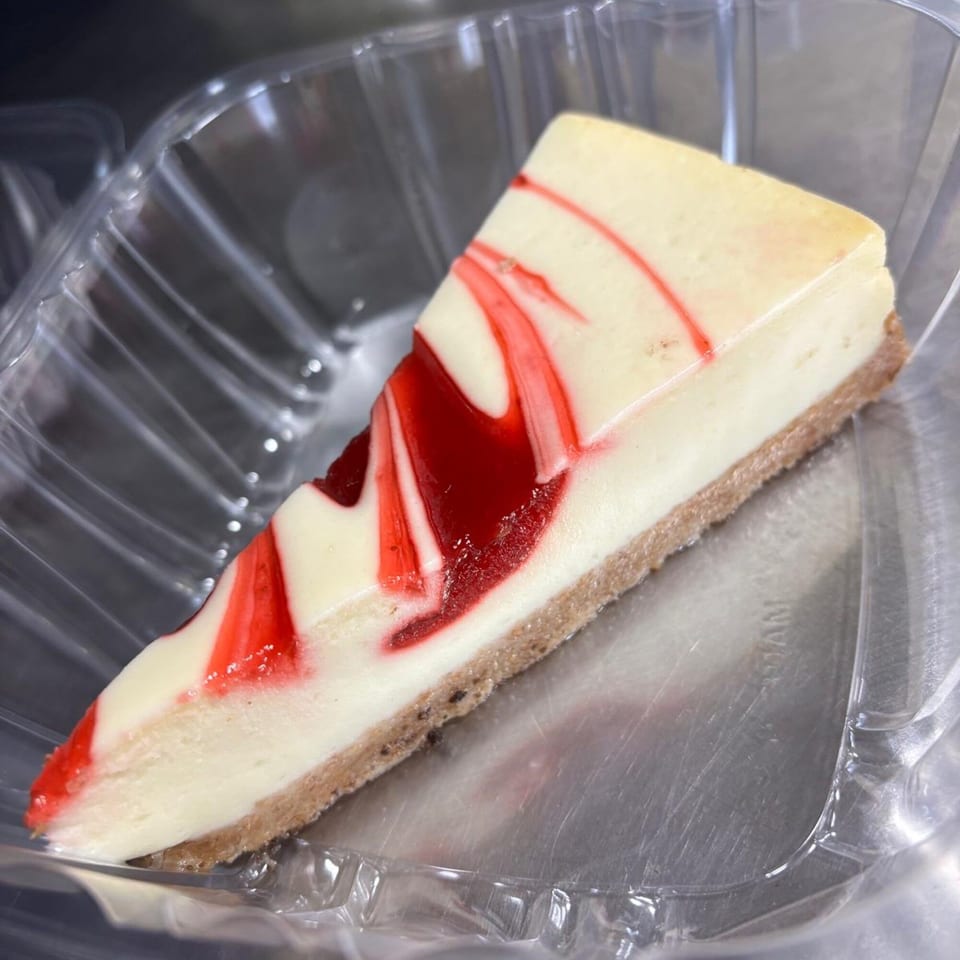 A slice of cheesecake with a graham cracker crust and red swirl topping in a plastic container.