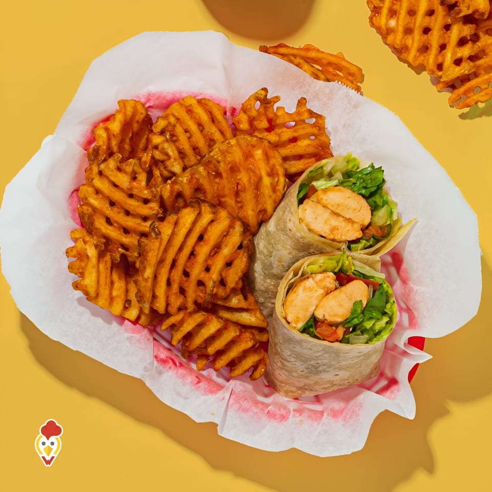 Two chicken wraps with lettuce and tomato, alongside a basket of waffle fries on a yellow background.